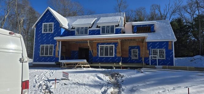 A two-story house under construction with blue weatherproofing wrap and snow covering the roof, yard, and driveway; trees are in the background.