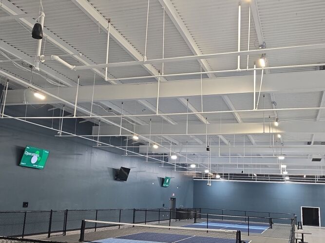 Indoor pickleball courts with blue nets, high white ceiling with hanging lights, and wall-mounted digital screens displaying information.