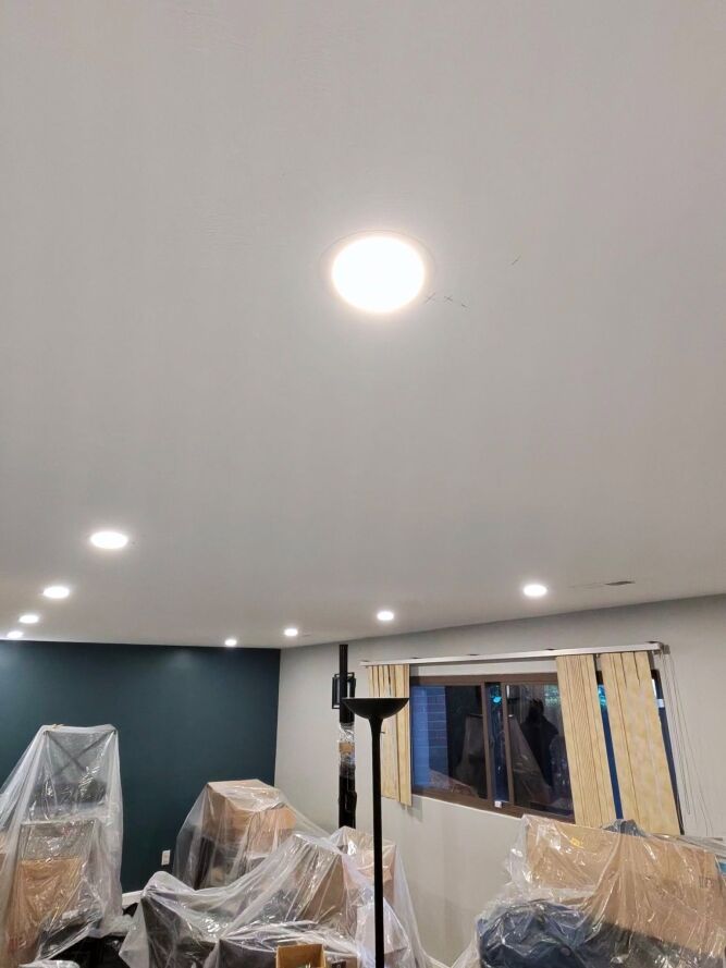 Ceiling with recessed lights on; room below contains furniture covered in plastic sheets, indicating ongoing painting or renovation work. A tripod stands near a window with partially covered blinds.