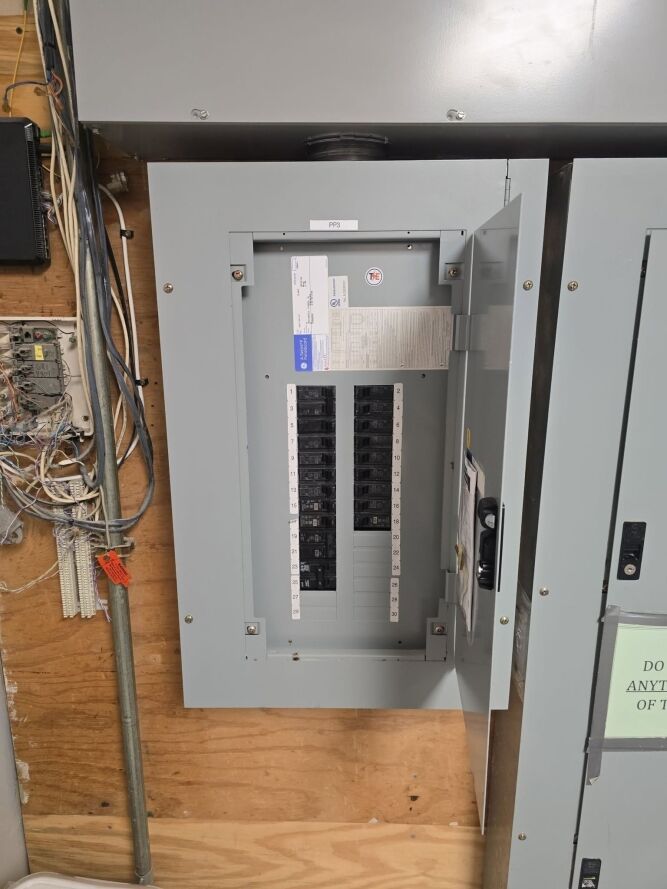 An electrical breaker panel with the door open showing circuit breakers and a label diagram, mounted on a plywood wall with visible wiring beside it.