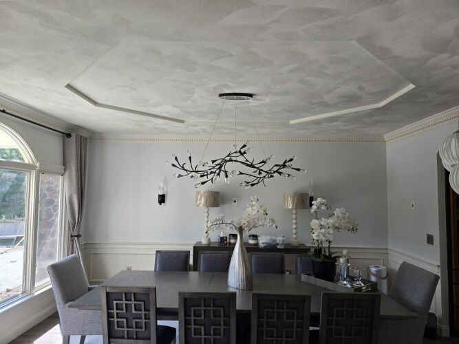 A modern dining room with a rectangular table, eight chairs, an abstract chandelier, decorative lamps, and white orchids on a sideboard under a textured ceiling.