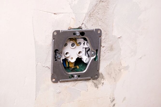 Exposed electrical wall socket with wires and mounting plate visible in an unfinished, unpainted wall.