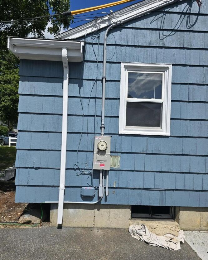 Blue house exterior with an electric meter and conduit mounted on the wall, a small window above, and a basement hatch partially covered by a cloth.