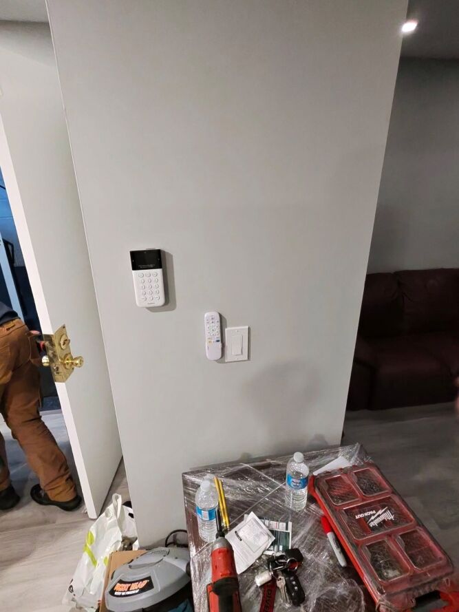 A wall with a security keypad, a remote, and a light switch; in front, a table with tools, water bottles, and plastic bags. A person is partially visible by an open door on the left.