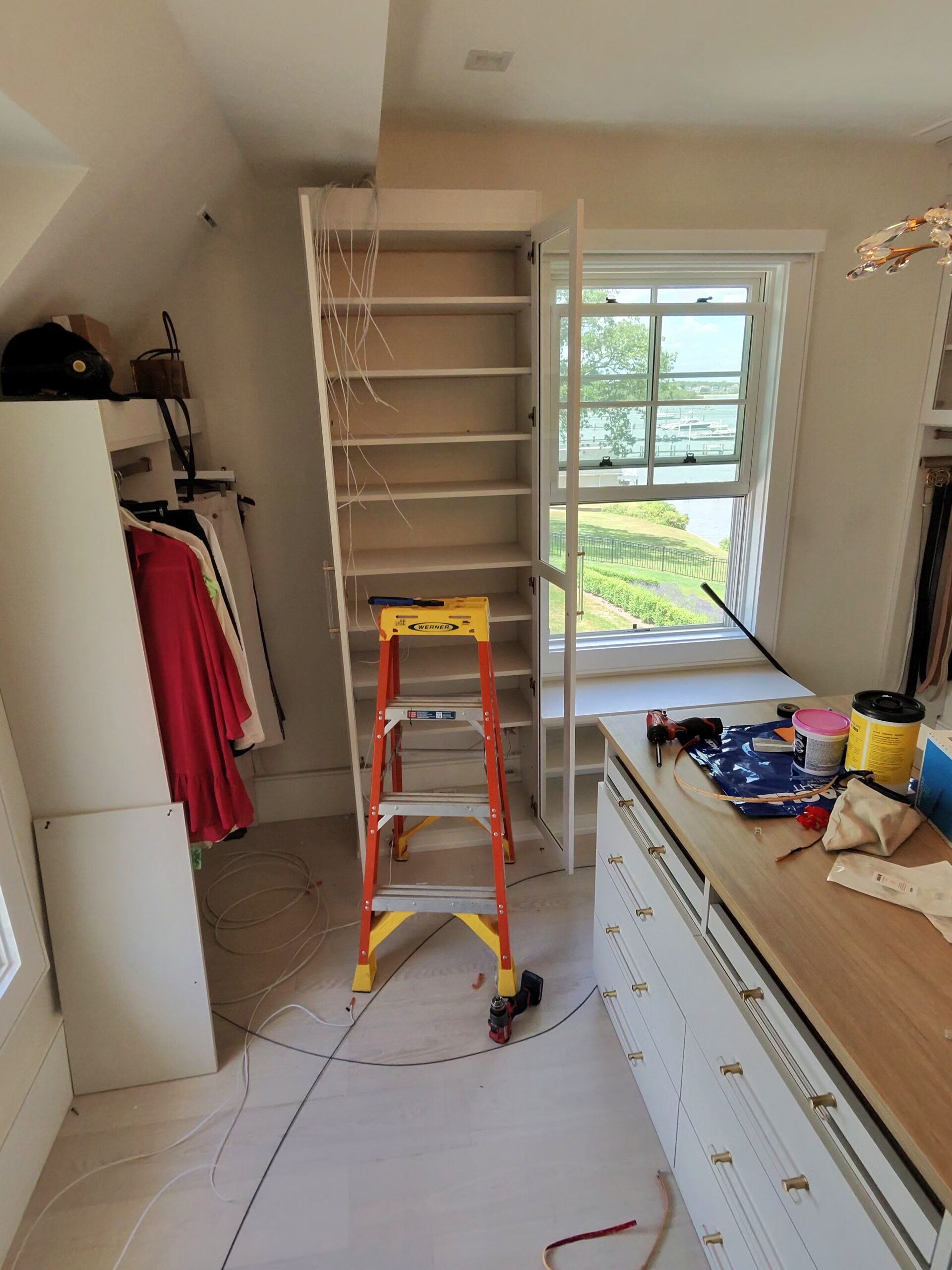 A step ladder stands in front of an empty bookshelf being assembled in a room with cabinets, tools, paint cans, and hanging clothes.