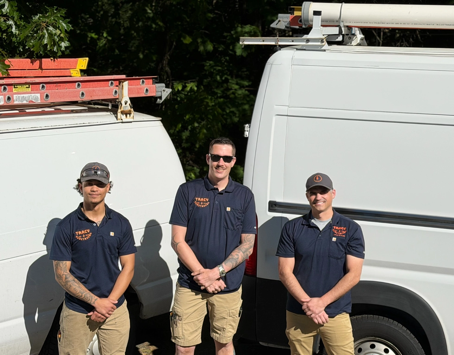 Three men in navy blue shirts and khaki shorts stand in front of two white work vans with ladders on top, posing outdoors in sunny weather.