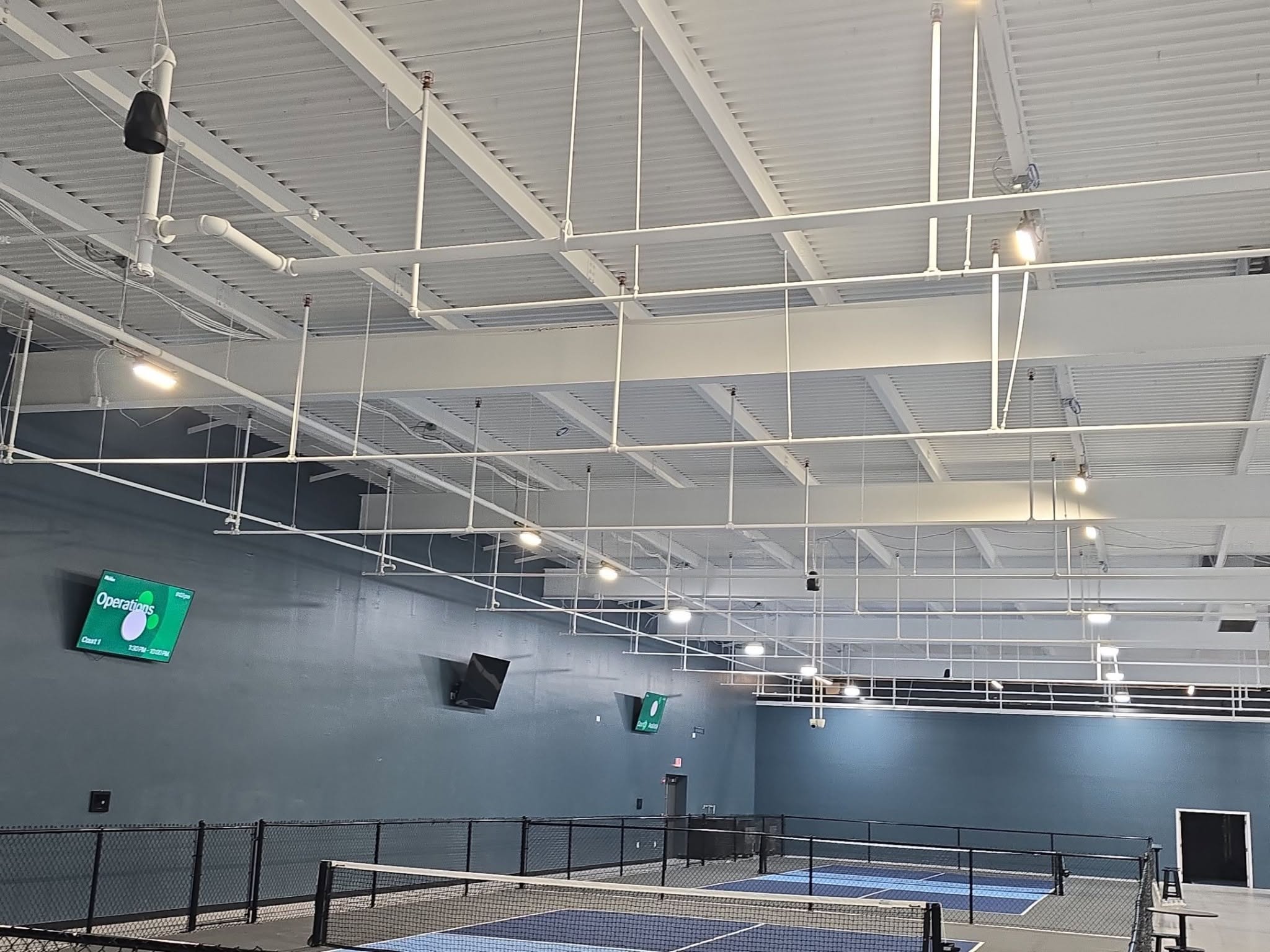 Indoor pickleball courts with blue nets, high white ceiling with hanging lights, and wall-mounted digital screens displaying information.