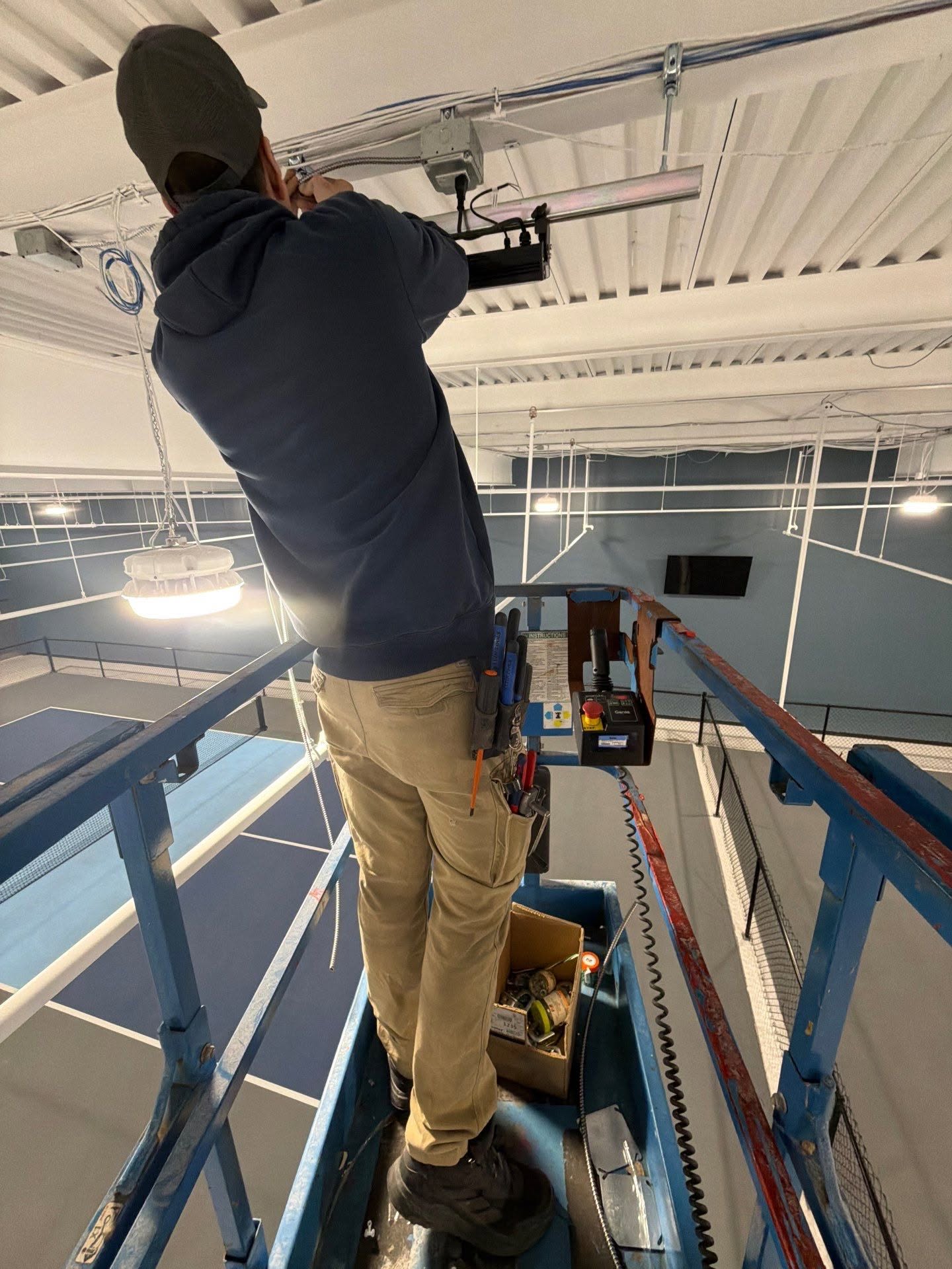 A person on a lift works on electrical wiring near the ceiling of an indoor sports facility, using various tools from a tool belt and box.