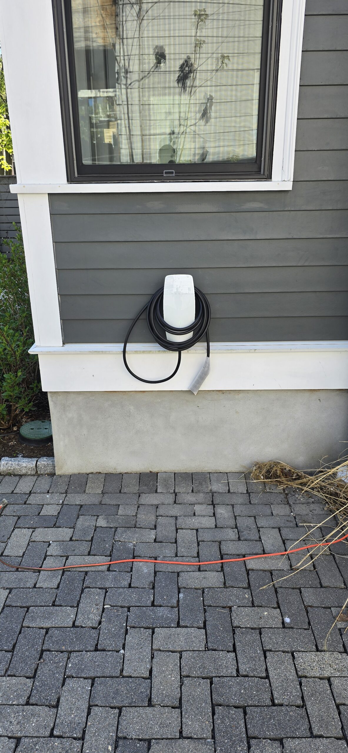 A white electric vehicle charging station with a coiled black cable is mounted on the side of a gray house above a brick driveway.