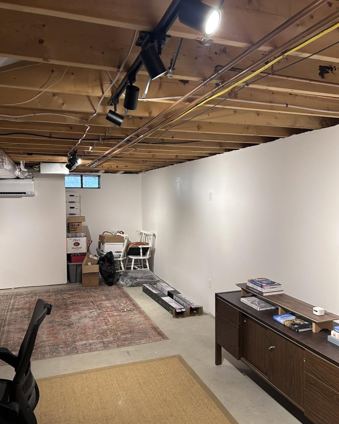 A basement room with exposed ceiling beams, white walls, office furniture, stacked boxes, chairs, and various items along the wall and floor.