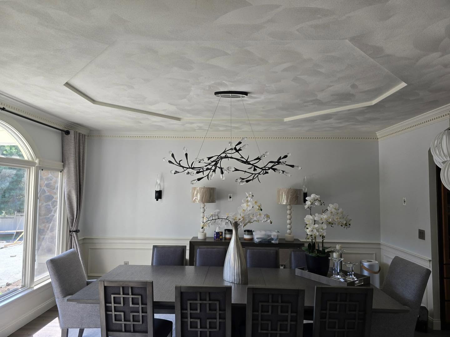 A modern dining room with a rectangular table, eight chairs, an abstract chandelier, decorative lamps, and white orchids on a sideboard under a textured ceiling.
