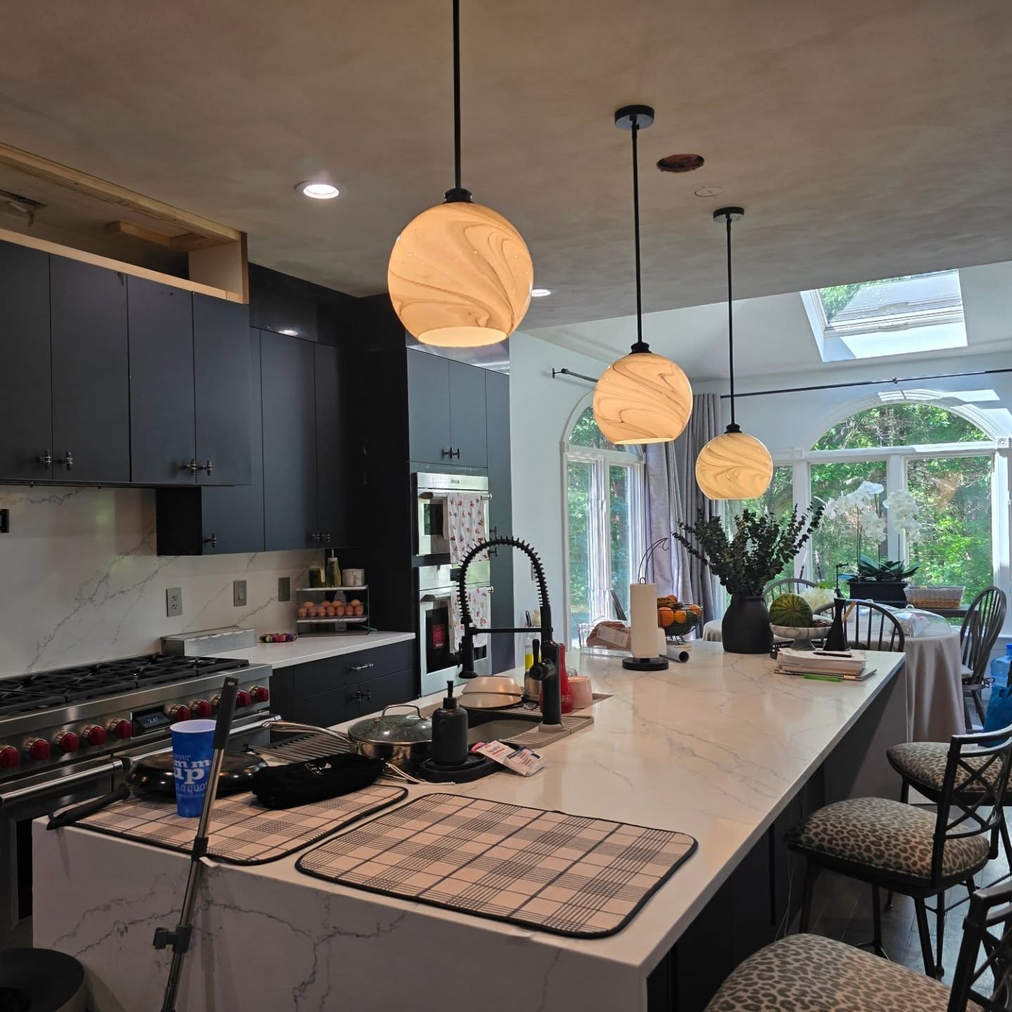 Modern kitchen with dark cabinets, marble countertops, pendant lights, and a large island with various items on top; a dining area with greenery is visible through the windows.