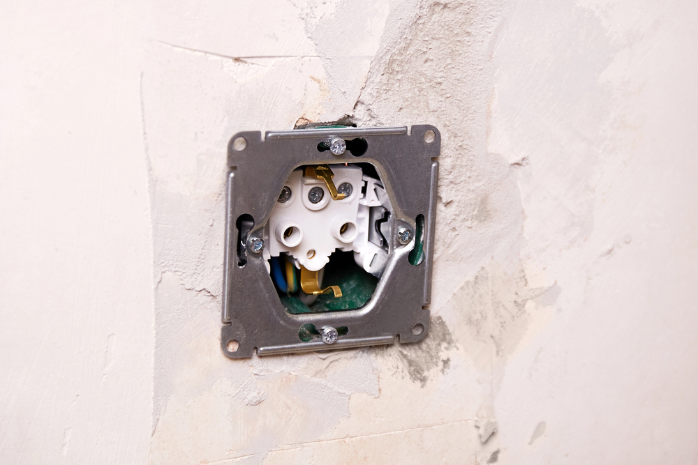 Exposed electrical wall socket with wires and mounting plate visible in an unfinished, unpainted wall.