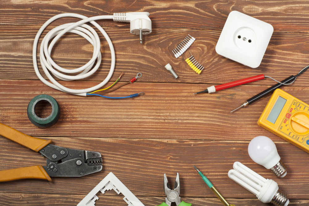 Various electrical tools and components, including wires, a plug, outlet, electrical tape, screwdriver, multimeter, light bulbs, and pliers, arranged on a wooden surface.