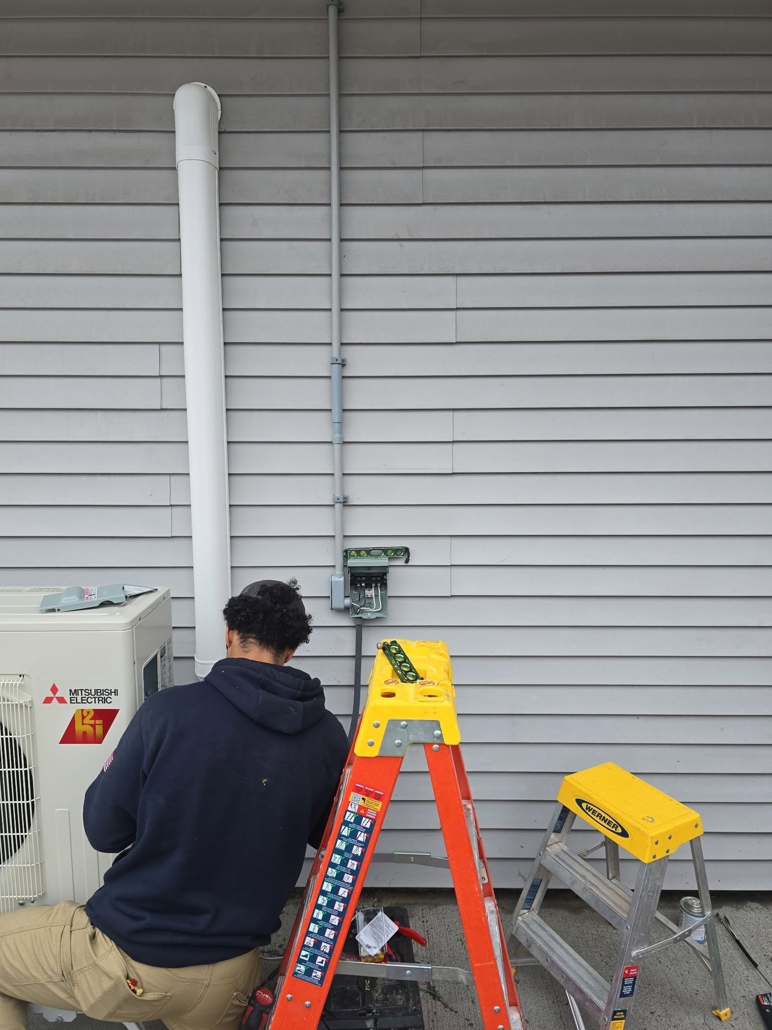 A person kneels near a Mitsubishi electric unit outside a building with gray siding, working on wiring beside two ladders and various tools.