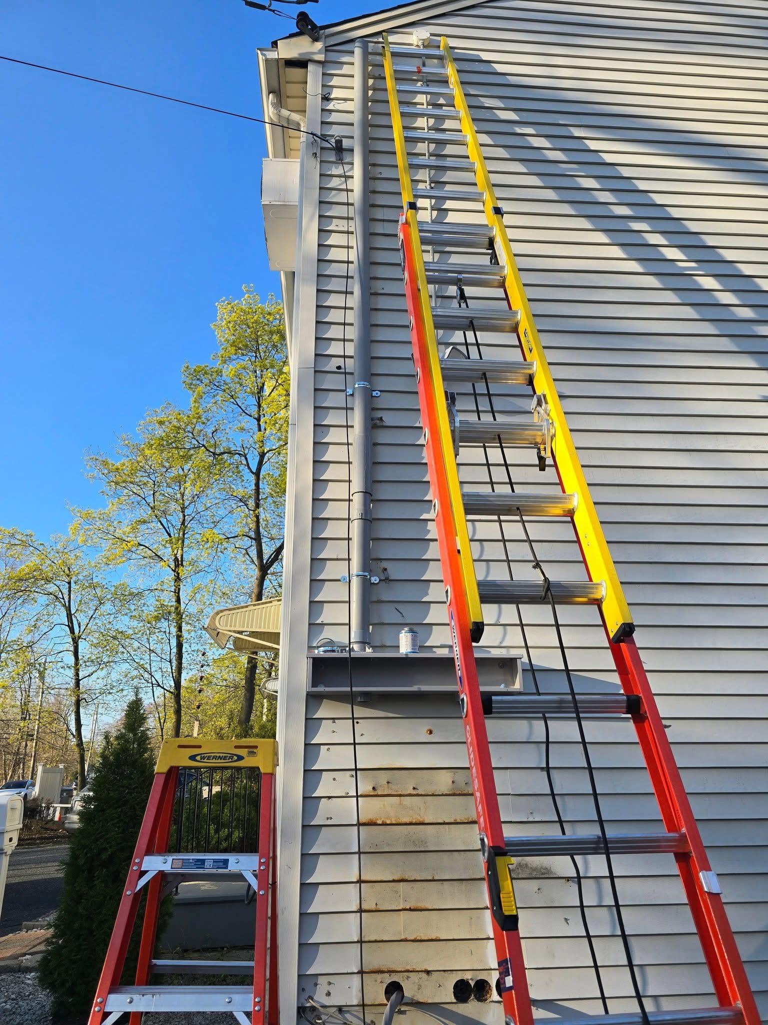 Two extension ladders are leaning against the side of a house with vinyl siding under clear blue skies. A step ladder is positioned nearby and equipment is attached to the siding.