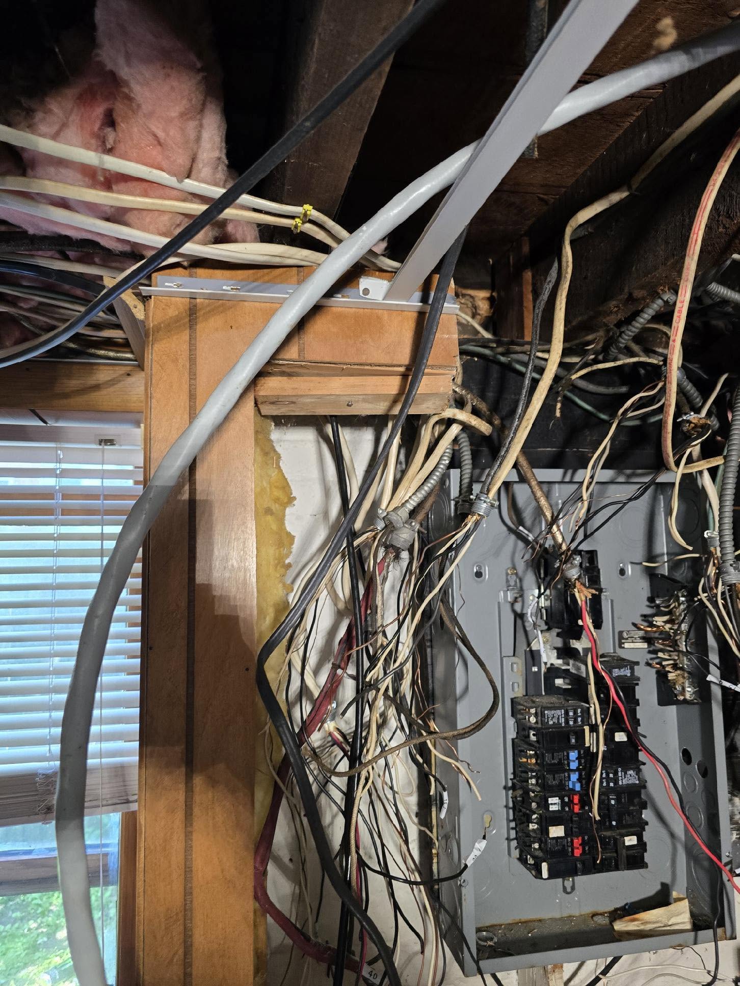 Electrical panel with exposed and tangled wires, some insulation, and wood framing visible; window with blinds on the left side of the image.