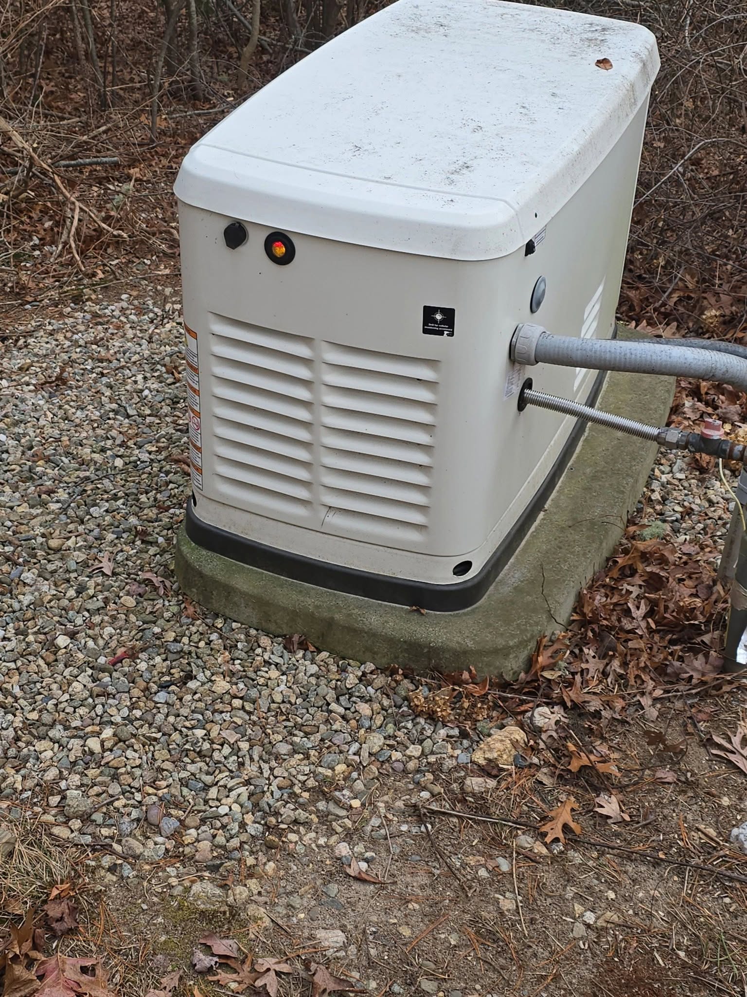 A standby generator sits on a concrete pad outdoors, surrounded by gravel and fallen leaves, with cables connected to its side.