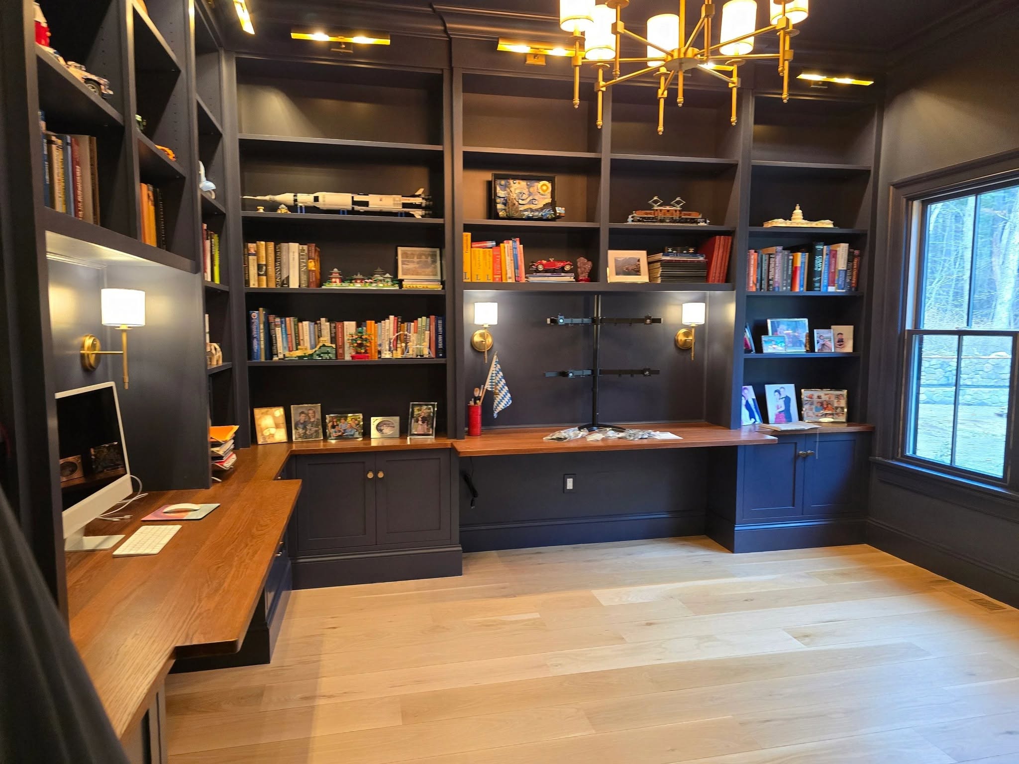 Modern home office with built-in dark wood bookshelves, a wooden desk with a computer, books, model cars, and framed photos, with a large window letting in natural light.