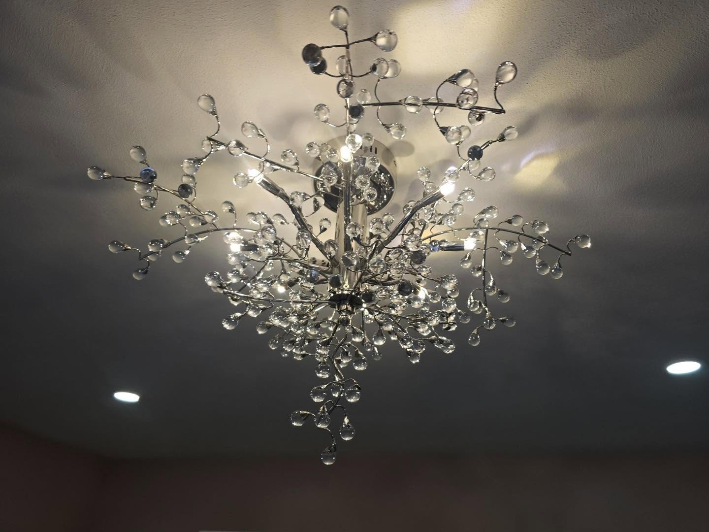 Modern crystal chandelier with branching metal arms and numerous hanging glass droplets, illuminated against a textured ceiling with recessed lights nearby.